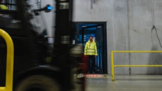 Un bărbat în echipament de protecție iese pe o ușă în hala unei fabrici. Un stivuitor trece pe lângă noi. Un bărbat în echipament de protecție iese pe o ușă în hala unei fabrici. Un stivuitor trece pe lângă noi.