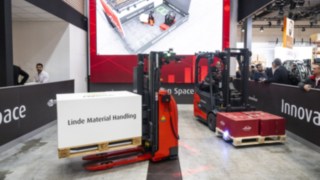 Două stivuitoare de la Linde MH transportă mărfuri. O imagine digitală a stivuitoarelor poate fi vizualizată pe un ecran.