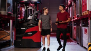 Două persoane în ținută sport se află în fața unui stivuitor cu catarg retractabil Linde MH Ri16; una dintre ele ține în mână o tabletă.